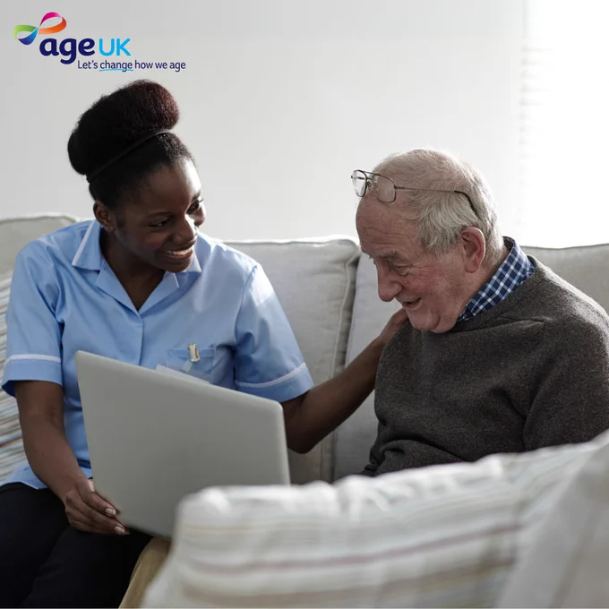 A healthcare worker, wearing a blue uniform, assists an older  individual with a laptop on a sofa. They are both smiling and engaged in conversation.