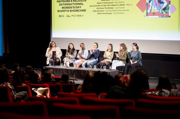 A panel of seven speakers sit on stage in front of an audience at a film screening event. They are seated behind a long table with microphones and water glasses. The backdrop displays event details: “Reclaim & Rejoice International Women’s Day Shorts Showcase,” along with a list of short film titles and filmmaker names. The atmosphere is warm and engaging, with the audience seated in red theater chairs, attentively watching the discussion.