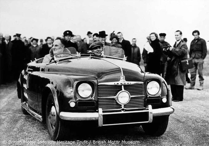 A black and white photograph of the Rover gas turbine car, JET1, with photographers, journalists and other people standing behind the car and looking on. The car is being driven by Maurice Wilks (Rover Company Technical Director). 