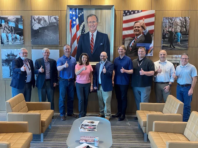 Nine people standing in front of the George H.W. Bush tribute wall at the George H.W. Bush Combat Development Complex.