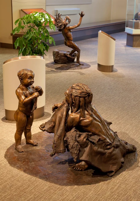 A museum display with a bronze sculpture with a squatting mother and standing child early human in front, and another bronze sculpture of a smaller early human behind 
