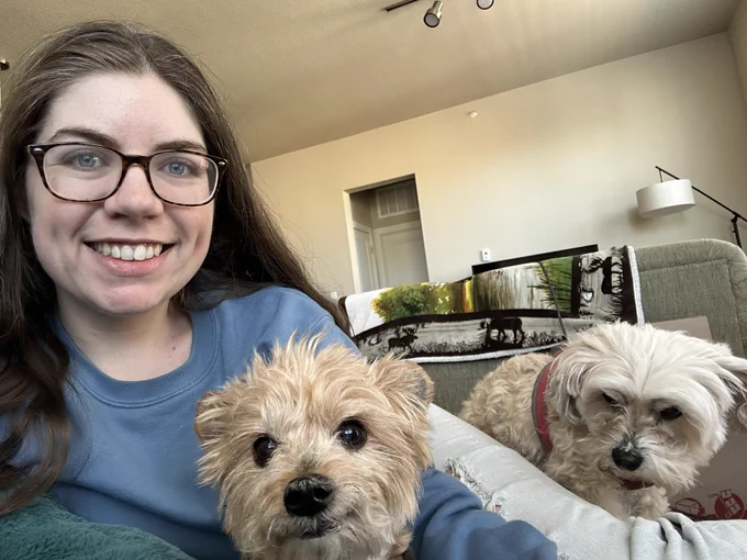Leah, a woman with long reddish/brown hair and blue eyes, is smiling towards the camera sitting beside her are two dogs, a small brown Yorkie/chihuahua mix and a white poodle/Yorkie mix.