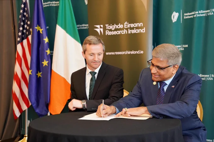 Two men in suits sit side by side to sign a document.