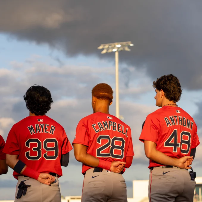 Marcelo Mayer, Kristian Campbell and Roman Anthony stand with their caps off during the National Anthem Thursday night during the Red Sox Spring Breakout game. They are all wearing red Red Sox uniforms with navy lettering.