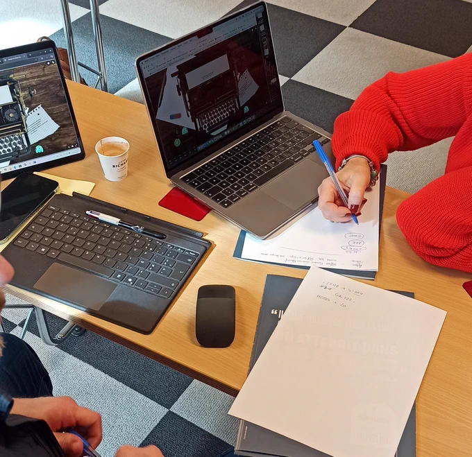 La photo montre une scène de travail collaboratif autour d’un projet numérique, probablement lié à un escape game ou à un dispositif pédagogique interactif.

🔹 Les écrans

Un ordinateur portable et une tablette affichent la même image d’une machine à écrire ancienne avec des feuilles de papier, ce qui suggère une interface numérique immersive ou un puzzle interactif.
Un second écran tactile avec un clavier détachable est également visible, renforçant l'idée d’un travail impliquant plusieurs supports numériques.
🔹 Les participants

Une personne portant un pull rouge écrit des notes sur une feuille avec un stylo bleu.
Une autre personne, en face, tient un stylo bleu, semblant engagée dans l’échange.
Seuls leurs mains et avant-bras sont visibles, mais l’interaction montre une dynamique de réflexion et de conception.
🔹 L’environnement

Un café dans un gobelet marqué "RICHARD" est posé sur la table, signe d’une séance de travail prolongée.
Une souris sans fil, un porte-cartes rouge, des 