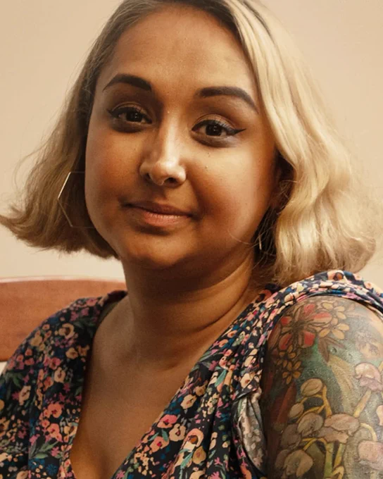 Sukhy looks at the camera towards her left shoulder. She is wearing a floral dress and has a tattooed arm nearest the camera.