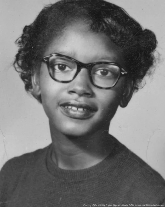 A black-and-white portrait of Claudette Colvin. She is wearing glasses and staring straight at the camera, lips parted in a half-smile.