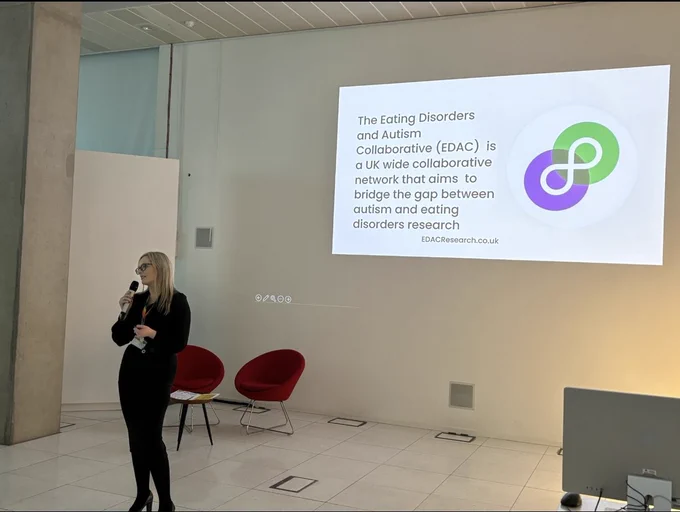 Woman standing with microphone in front of a slide in the background that says ‘The Eating Disorders and Autism Collaborative (EDAC) is a UK wide collaborative network that aims to bridge the gap between autism and eating disorders research. EDACResearch.co.uk’ with the symbol for EDAC (which is a purple and green circle interlinking around a white neurodiversity symbol).  Behind the woman is two seats and a table.