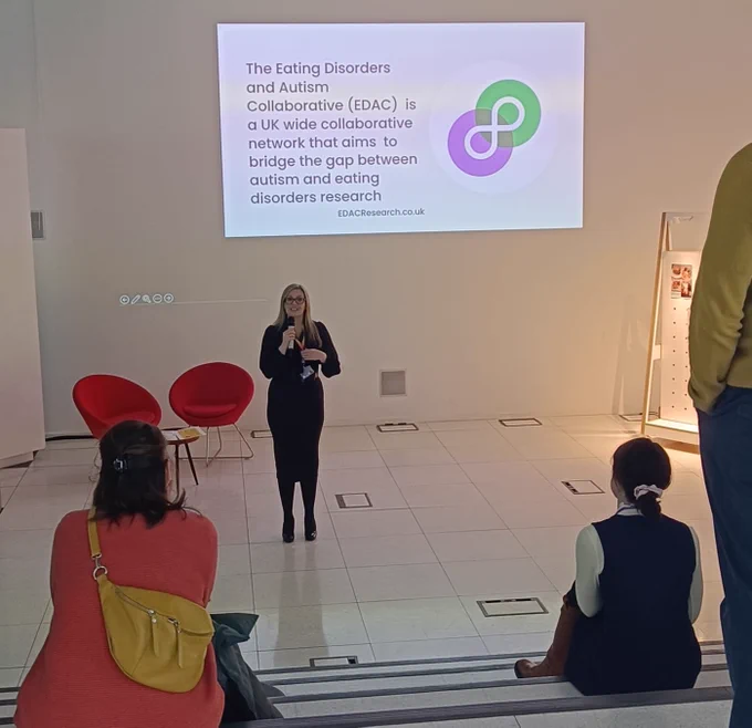 Woman standing with microphone in front of some seated guests with a slide in the background that says ‘The Eating Disorders and Autism Collaborative (EDAC) is a UK wide collaborative network that aims to bridge the gap between autism and eating disorders research. EDACResearch.co.uk’ with the symbol for EDAC (which is a purple and green circle interlinking around a white neurodiversity symbol).  Behind the woman is two seats and a table.