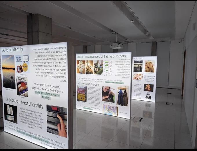 A wide open space with 3 frames which have canvases that are lit up.  The canvases have a selection of images and text outlining the experiences of Autistic people with Eating Disorders.  There is a projector above the canvases.  