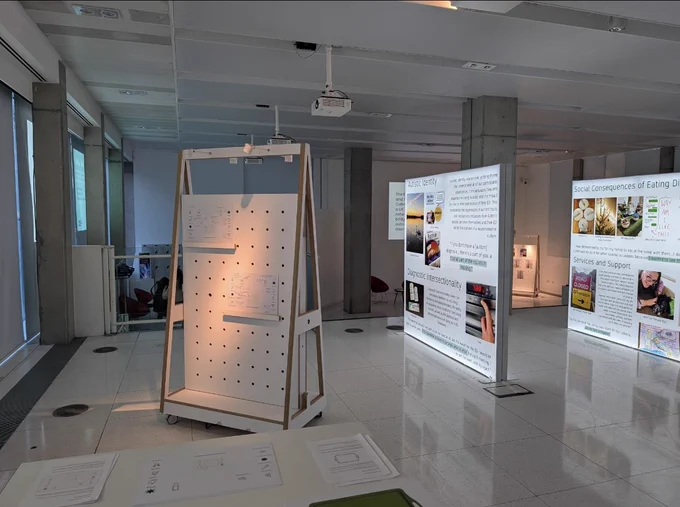 A wide open space with 2 frames which have canvases that are lit up.  The canvases have a selection of images and text outlining the experiences of Autistic people with Eating Disorders.  There is a projector above the canvases.  A stand displaying menu sheets that detail how eating environments can be improved for Autistic people.  In front of a stand is a white table with a blank eating environment sheet and instructions for visitors to have a go at designing autism affirming eating environments.  Behind one of the canvases is a big grey column and behind that is a stand with images displayed representing the experiences of an Autistic person with Intellectual Disability who also had an eating disorder.  There is a row of projectors above the stands and canvases.  

