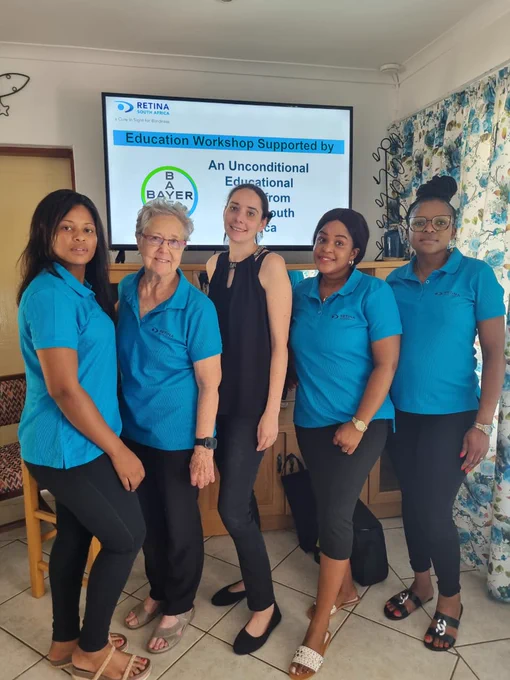 he team is gathered for a photo in front of a screen showcasing details about an "Education Workshop Supported by Bayer" and "Retina South Africa." Judy from the Margate Old Age Home stands prominently in the centre of the image.