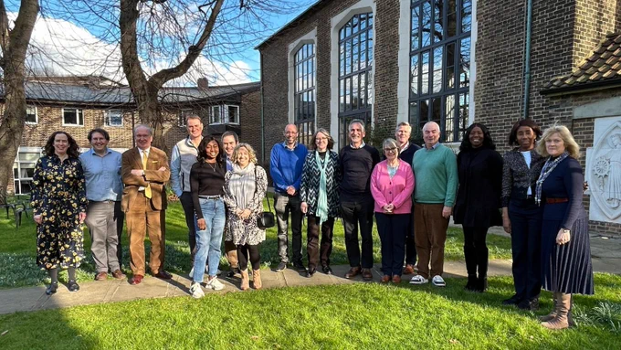 Sixteen people standing outside in the sunlight, on a cemented pathway, beside trees and a grass lawn.