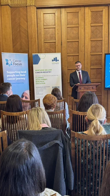 Colin speaks in front of a room of seated people. There are vinyl popup banners beside him for Cancer Focus NI and the NI Cancer Registry