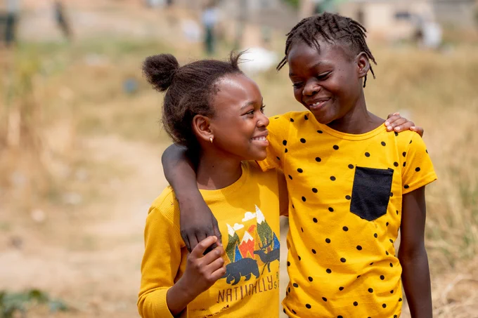 Photo shows two young girls in matching yellow outfits smile at each other while walking outdoors, with one girl’s arm around the other in a warm gesture of friendship.