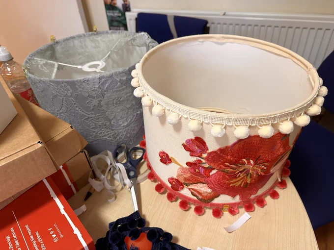 Two hand-decorated lampshades.  One is a grey-green lace. One is cream with bright red large flowers.