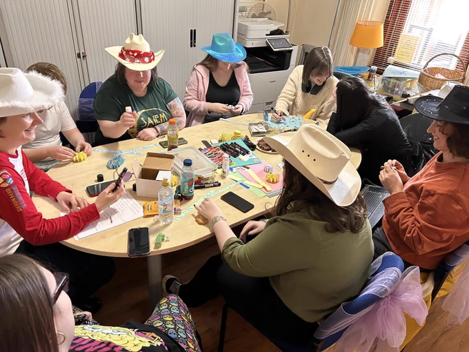 A group of adults and teenagers sit round a table. They are all wearing different coloured cowboy hats. There are lots of crafting resources in the middle of the table such as paper, pens and glue.  They all look quite happy focusing on their activity.