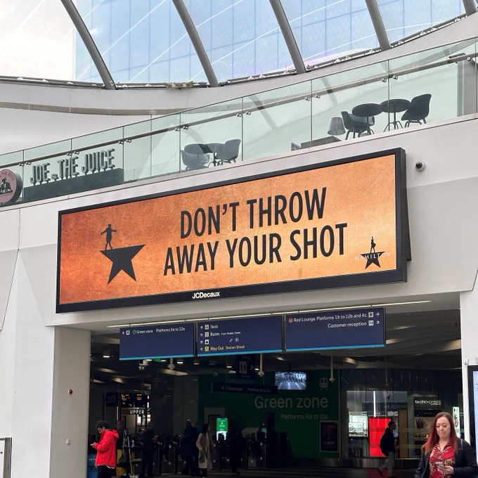 Digital sign at a train station displaying the message "DON'T THROW AWAY YOUR SHOT" with a star logo of Hamilton.