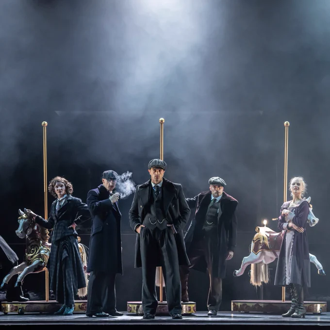 A scene from a theatrical dance production of Peaky Blinders featuring five individuals in period costumes on a dimly lit stage, with carousel horses behind them.