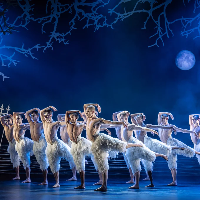 A group of ballet dancers from Matthew Bourne's New Adventures perform in "Swan Lake," wearing feathered costumes with a moon in the backdrop.