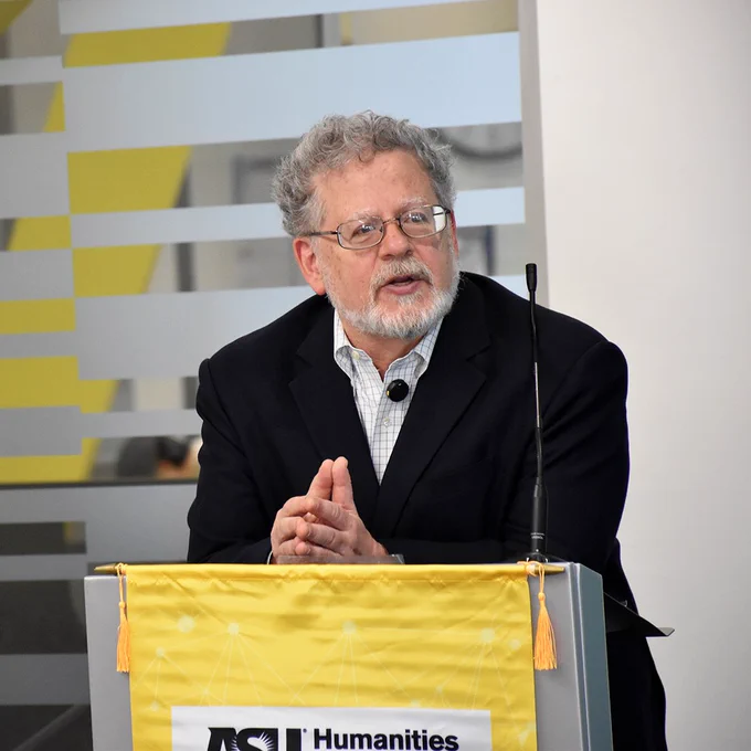 Leonard Cassuto speaks during his presentation. His hands are clasped together and he is leaning against a podium, which has a yellow cloth with the ASU Humanities Institute logo draped over it.