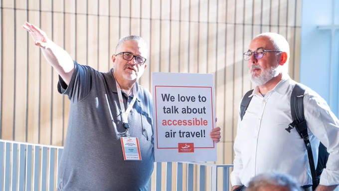 Two males at the 2024 All Wheel’s Up Global Forum in conversation while the male on the left of the image holds a white sign with black and orange writing reading, “We love to talk about accessible air travel!”. The AWU logo in orange with white writing is at the bottom of the sign.