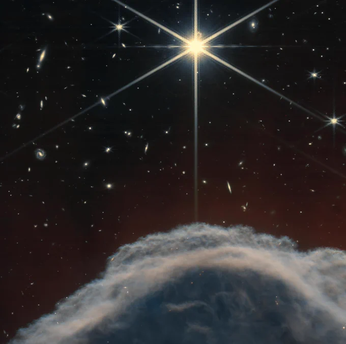 A close-up view of the Horsehead Nebula's edge captured by NIRCam, revealing the intricate texture of the cloud's upper layer. A central bright star, with its characteristic eight-pointed diffraction spikes, dominates the image. Faint, distant galaxies are scattered throughout the deep space background, which has a reddish tint.