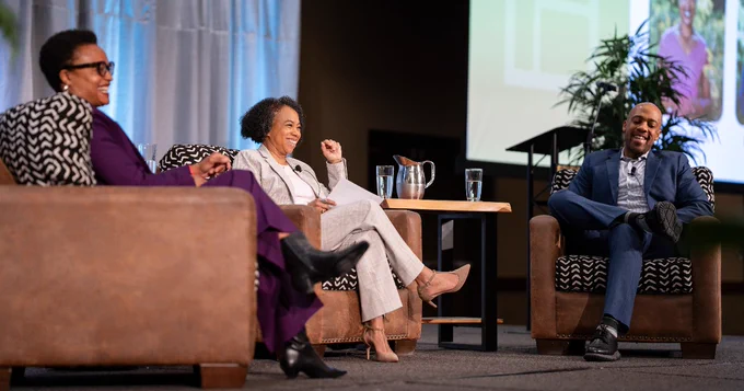 Shalanda Baker, Sandra Henry, and Mandela Barnes laughing and engaging in conversation on stage at a conference, seated comfortably with a large screen displaying a presentation in the background.