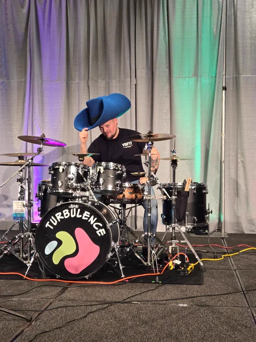 A musician playing a drum set labeled "Turbulence" on stage, surrounded by various microphones and cables.