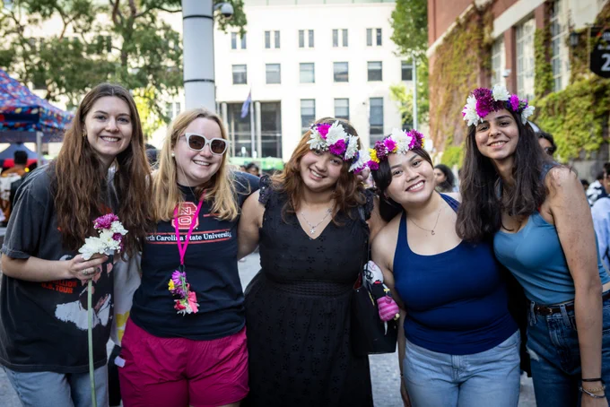 Five students pose and smile for the camera, 3 on the right are wearing bright pink and white flower crowns