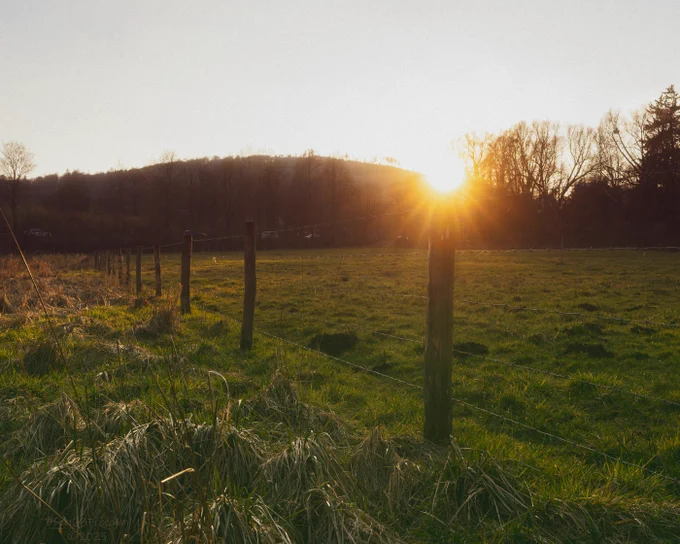A tranquil rural landscape at sunset, featuring a grassy field bordered by a wooden barbed wire fence and bathed in warm golden light.