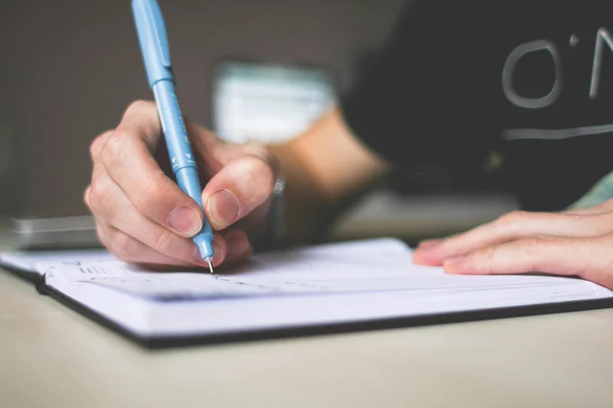 A zoomed in shot of a person sitting at a desk writing on a notebook with a pen.