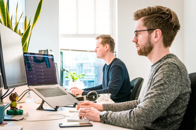 Zwei junge Männer in einem hellen, lichtdurchfluteten Raum am Schreibtisch sitzend, arbeiten am PC. Der Mann im Vordergrund trägz Brille und Bart, der im Hintergrund ist glattrasiert ohne Brille.