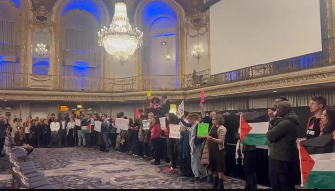 dozens of protesters in a ballroom holding signs condemning genocide, scholasticide and censorship 