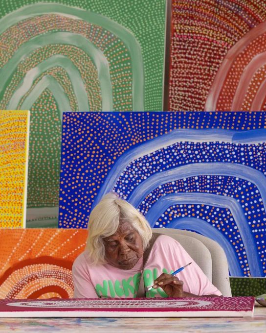 [ID] Yindjibarndi artist Alice Guiness painting on a canvas, with a sample collection displayed in the background. 
