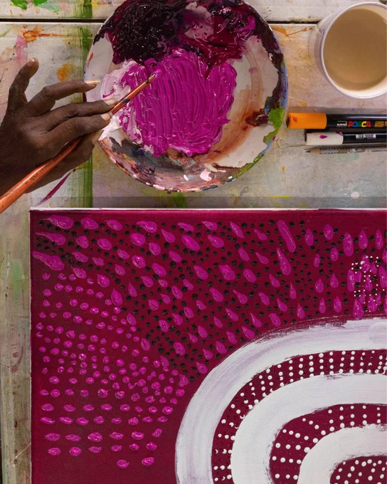 [ID] An artwork in progress, with a pot of paint beside it and a hand dipping a paintbrush into the pot. 
