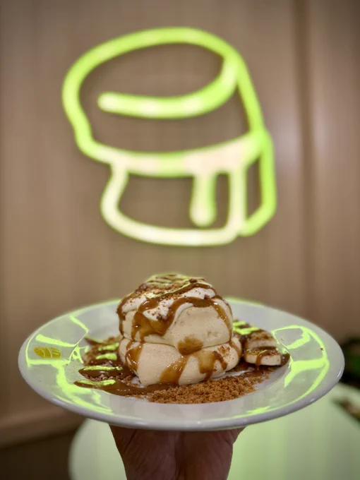 A person holding a plate with a caramel-topped dessert, with a neon sign of the Fluffy Fluffy logo in the background.