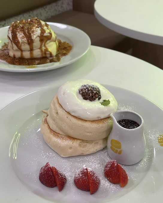 A photo of two plates of fluffy pancakes at a café. The front plate features pancakes topped with a dollop of cream, a chocolate ball, and served with strawberries and a small jar of sauce. The rear plate has pancakes drizzled with caramel, served on a bed of crumbs.