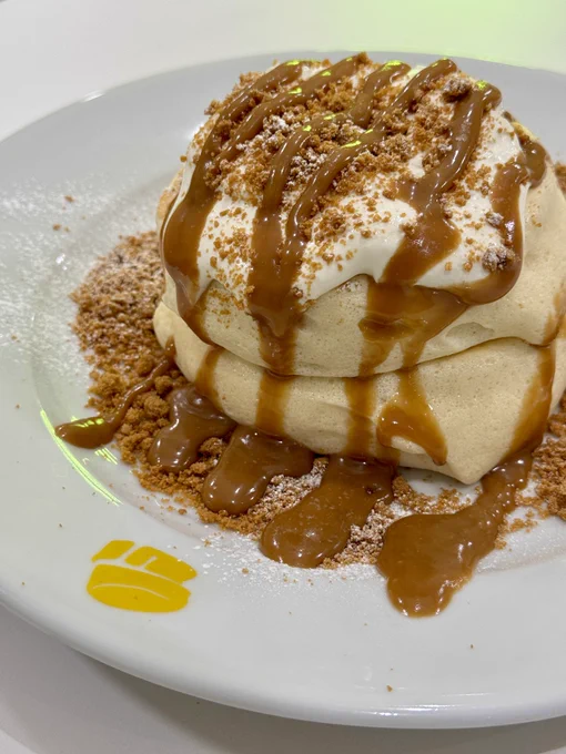 A plate of stacked pancakes topped with whipped cream, drizzled with caramel sauce, and sprinkled with crushed biscuits, served at Fluffy Fluffy, indicated by a small yellow logo on the plate.