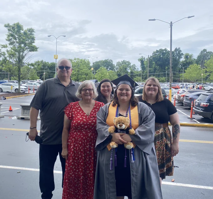 Group of five individuals posing for a photo in a parking lot on a cloudy day, celebrating a graduation. The graduate is holding a plush bear and wearing a cap and gown.