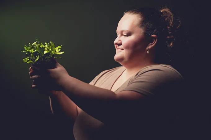 A person smiling as they hold a small potted plant with lush green leaves, illuminated by soft light against a dark background.