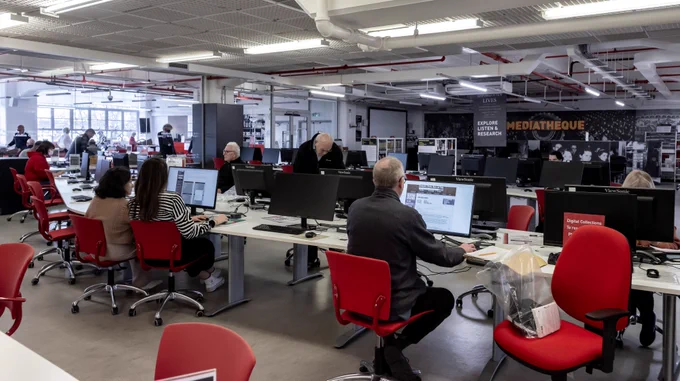 A large group of people conducting online research in an information area. Behind rows of desks with computer monitors, there is a for a Mediatheque.