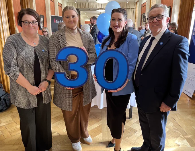 Image of four people holding branding marking 30 years of National Lottery funding – Kate Beggs, NI director of the National Lottery Community Fund; Junior Minister Aisling Reilly; Deputy First Minister Emma Little-Pengelly and Justin King, Chair of lottery operator Allwyn UK.