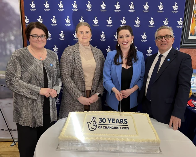 Image of four people cutting a cake decorated with the National Lottery logo and wording ’30 years of changing lives’ – Kate Beggs, NI director of the National Lottery Community Fund; Junior Minister Aisling Reilly; Deputy First Minister Emma Little-Pengelly and Justin King, Chair of lottery operator Allwyn UK.