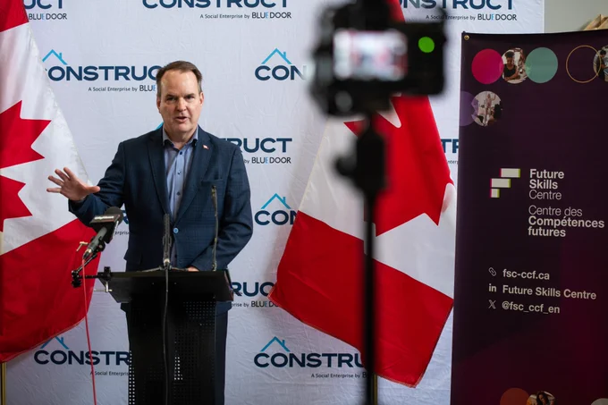 The Honorable Steven MacKinnon, Minister of Employment, Workforce Development and Labour speaking at a podium with two Canadian flags in the background.