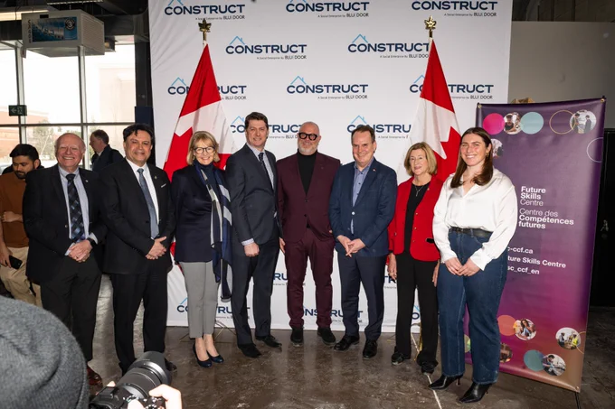 Local MPs, Tony van Bynen, Majid Jowhari, Leah Taylor Roy, and Helena Jaczek, FSC's Executive Director, Noel Baldwin, the Honorable Steven MacKinnon, Minister of Employment, Workforce Development and Labour, Blue Door's CEO Michael Braithwaite and program lead Emma Wood standing, smiling in front of two Canadian flags at Blue Door's training centre. 