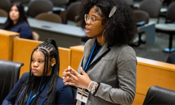 Two students in a mock courtroom. One stands and speaks and the other sits and listens.