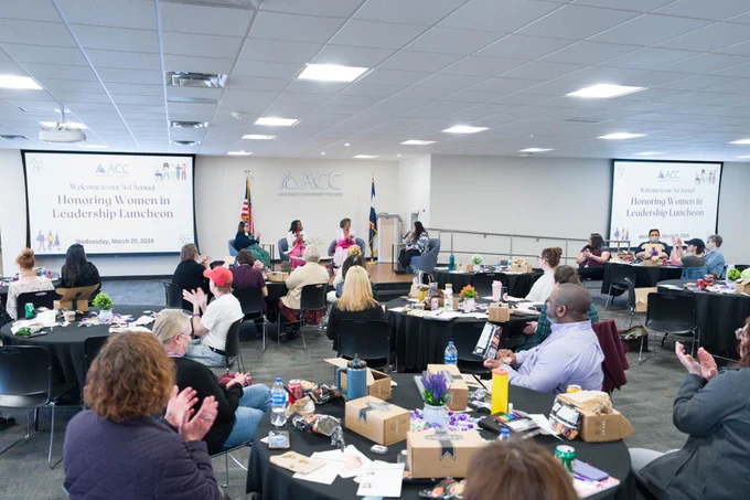 A photograph of the 2024 Honoring Women in Leadership Luncheon in Arapahoe Community College's Summit Room. The room is full of round tables with guests sitting and looking a the stage where four panelists are giving a talk. 