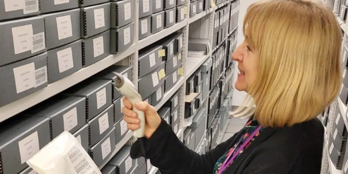 Curator, Janette Martin, standing in front of neatly arranged archive boxes on shelves holding a barcode scanner.