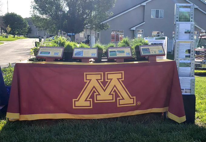 A display table set up on a lawn, with educational signs, brochures, and turfgrass and weed samples in pots. The table cloth shows the University of Minnesota "M" logo. 